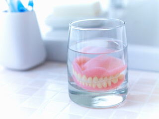 Set of dentures in a glass of fluid on a white tile counter top.